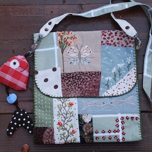 May include: A patchwork shoulder bag with a whimsical design. The bag features various fabric squares with floral, butterfly, and fish motifs. A red plaid fish-shaped charm hangs from the bag. The bag has a long, light green strap.