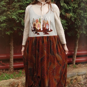 May include: A woman wearing a long, brown and orange patterned dress with a white crocheted hat and a light blue sleeveless top with an embroidered forest scene.