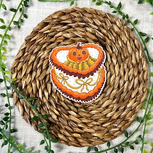 Puede incluir: Una pegatina en forma de corazón con una cara de calabaza naranja central con un tallo marrón oscuro, rodeada por capas de glaseado naranja y marrón, decorada con pequeños lazos amarillos y una textura de puntos sobre fondo blanco.