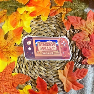 Puede incluir: Una pegatina de una consola de videojuegos con una pantalla que muestra el texto "Stardew Valley". La consola es rosa y tiene un fondo de madera marrón. La pegatina está sobre un fondo tejido con hojas de otoño.