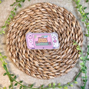 Puede incluir: Pegatina de consola de juegos Nintendo Switch con tem&aacute;tica de Animal Crossing, rosa y blanca. La pegatina presenta una consola de juegos rosa y blanca con una pantalla que muestra el logotipo de Animal Crossing y una escena de bosque verde.