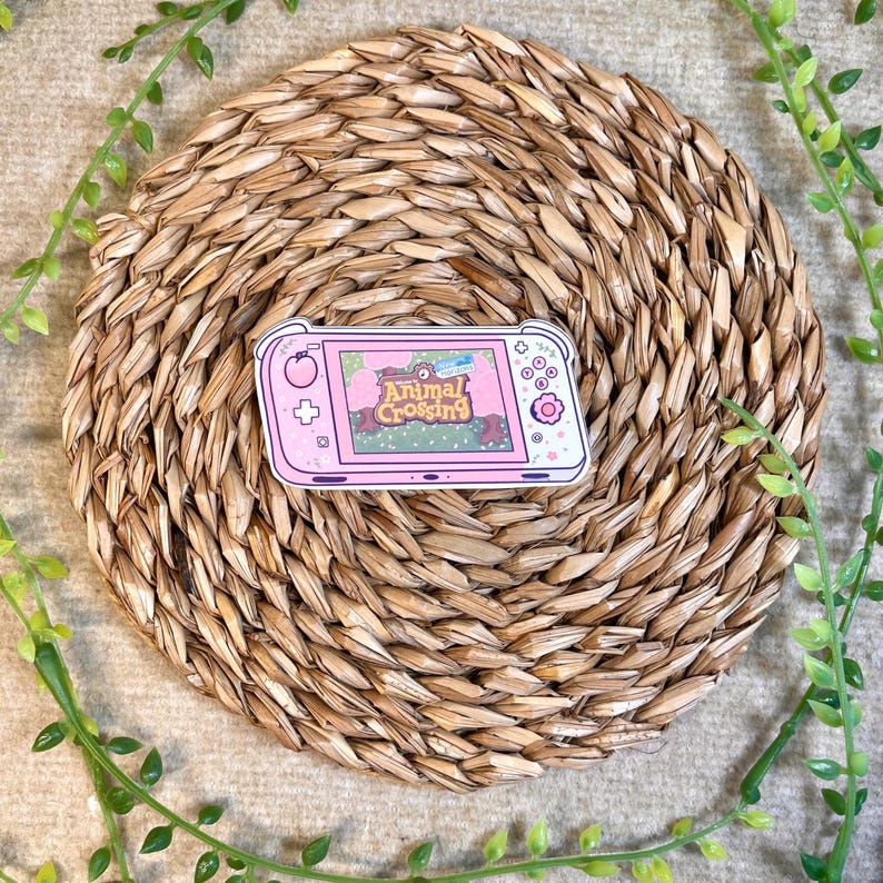 Puede incluir: Pegatina de consola de juegos Nintendo Switch con tem&aacute;tica de Animal Crossing, rosa y blanca. La pegatina presenta una consola de juegos rosa y blanca con una pantalla que muestra el logotipo de Animal Crossing y una escena de bosque verde.