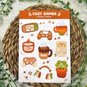 May include: A sticker sheet titled COZY GAMER by Wistaria.blooms featuring illustrations of a handheld console, game controller, steaming mugs, a candle labeled cozy homebody, a game console with Game Over text, headphones, and a potted plant.