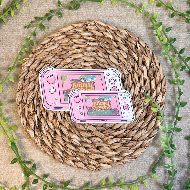 Puede incluir: Dos pegatinas de consola de juego Nintendo Switch Animal Crossing de color rosa y blanco. Las pegatinas presentan el logotipo de Animal Crossing y un fondo verde y marr&oacute;n.