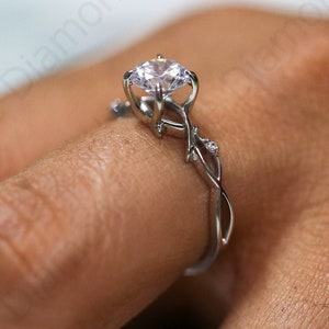Könnte beinhalten: Ein silberner Ring mit einem großen, klaren Edelstein. Der Ring hat ein zartes, verschlungenes Rankenmuster mit kleinen Akzentsteinen. Der Ring befindet sich am Finger einer Person und zeigt die filigranen Details und das Funkeln des Schmucks.