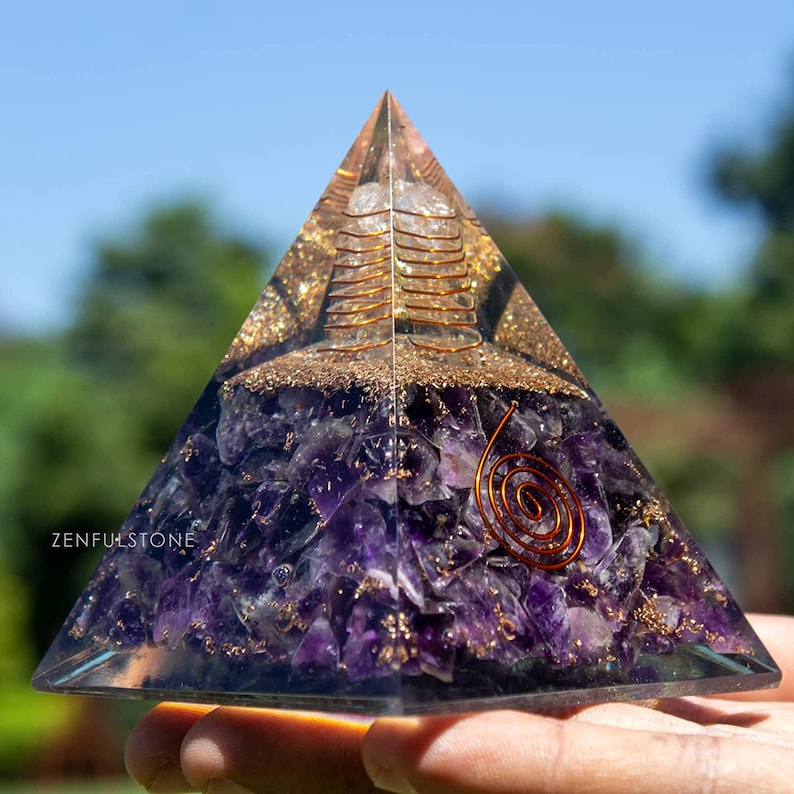 May include: A clear crystal pyramid with a copper wire spiral inside. The pyramid is filled with amethyst crystals and gold glitter. The pyramid is being held in a hand.