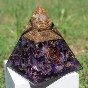 May include: A clear crystal pyramid with a copper wire spiral inside. The pyramid is filled with amethyst crystals and gold glitter. The pyramid is sitting on a white surface. "Zenfulstone" is written on the surface.