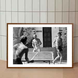 May include: Black and white photograph of three men playing table tennis outdoors. The men are wearing shorts and are shirtless. The table tennis table is in the foreground, and there is a building in the background.