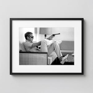 May include: Black and white framed print featuring a man in sunglasses holding a gun while seated. The man is wearing a light-colored shirt and pants, and white sneakers. The print is displayed in a black frame.