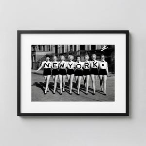May include: Black and white framed print featuring a group of women in matching outfits, posing outdoors. The word "NEW YORK" is spelled out in large, bold letters above them. The women are wearing black shorts, white tops, and black heels.