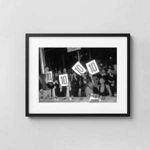 Könnte beinhalten: Ein schwarz-weiß gerahmtes Foto, das eine Gruppe von Menschen an einem Tisch zeigt, die weiße Schilder mit der aufgedruckten Zahl 10 hochhalten. Die Szene fängt eine feierliche Atmosphäre ein, in der Personen ihre Wertungskarten heben.