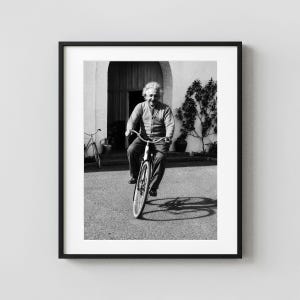 May include: A black and white framed photograph featuring Albert Einstein riding a bicycle. He is wearing a cardigan and trousers, positioned in front of an arched doorway. A second bicycle is visible in the background near a small tree.