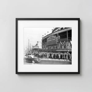 Puede incluir: Una fotografía enmarcada en blanco y negro de Wrigley Field, sede de los Chicago Cubs, con una gran multitud reunida frente a la entrada del estadio. Se ve un coche antiguo en primer plano y banderines colgados en el exterior del estadio.