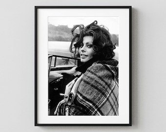 May include: A black and white framed photograph featuring a person with voluminous dark hair looking over their shoulder. The individual is wearing a plaid coat with a dark collar, positioned next to the open door of a vintage car.