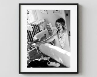 May include: A black and white framed photograph of a person sitting in a bathtub, wearing a button-down shirt, reading a book titled Help Your Husband Get Ahead, with a single white sneaker resting on the floor near the edge of the tub.