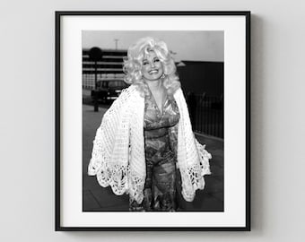 May include: A black and white framed photograph featuring a smiling individual with voluminous blonde hair, wearing a patterned jumpsuit and a large, intricate white crochet shawl draped over the shoulders, standing outdoors.
