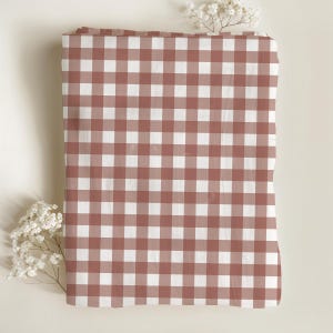May include: A rectangular fabric piece featuring a classic gingham pattern in muted terracotta red and white. The textile displays a consistent grid of squares across its surface, accented by small sprigs of white baby's breath flowers.