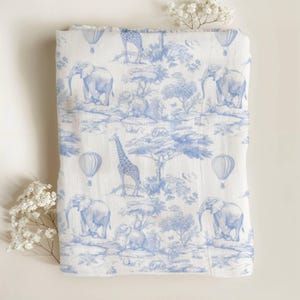 May include: A folded white and blue muslin blanket featuring a toile design with elephants, giraffes, hot air balloons, and trees. The blanket has a soft, lightweight appearance, suitable for a baby or child.