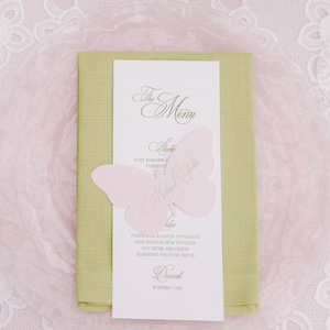 May include: A white rectangular menu card with green text titled The Menu, featuring a light pink butterfly-shaped paper cutout overlay. The menu is placed on a folded light green napkin, which rests on a clear glass plate with a scalloped edge.