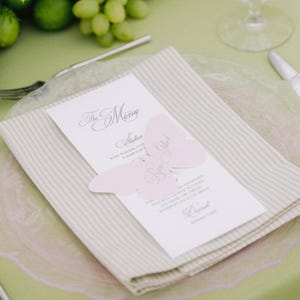 May include: A white rectangular menu card with black text titled The Menu, featuring a light pink butterfly-shaped paper cutout placed over the center. The menu rests on a folded beige and white striped napkin atop a clear glass plate.