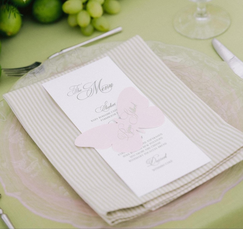 May include: A white rectangular menu card with black text titled The Menu, featuring a light pink butterfly-shaped paper cutout placed over the center. The menu rests on a folded beige and white striped napkin atop a clear glass plate.