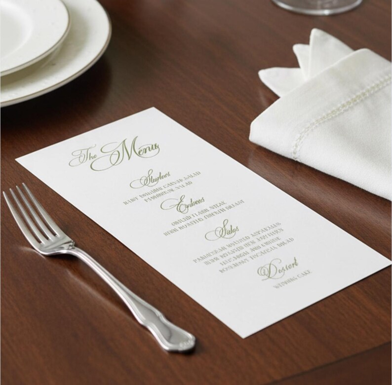 May include: A white rectangular menu card with elegant green cursive text featuring sections for Starters, Entrees, Sides, and Dessert. The menu is positioned next to a silver fork and a folded white cloth napkin on a dark wooden surface.