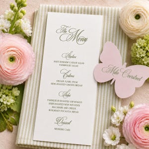 May include: A vertical white menu card with dark green text titled The Menu, listing starters, entrees, sides, and dessert. A light pink butterfly-shaped place card with the name Mike Overstreet is placed next to the menu on a striped fabric.