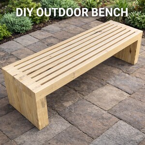 May include: A light-coloured wooden outdoor bench with a slatted seat and block legs. The bench is made of unfinished wood and is set on a brick patio. The text "DIY OUTDOOR BENCH" is at the top of the image.