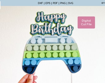 May include: A blue, green, and white pop-it shaped like a video game controller with the words "Happy Birthday" above it.