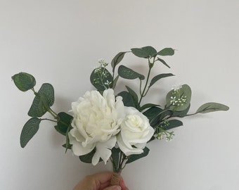 May include: A small bouquet of white flowers with green leaves, tied with twine. The bouquet includes a white peony, a white rose, and small white flowers.