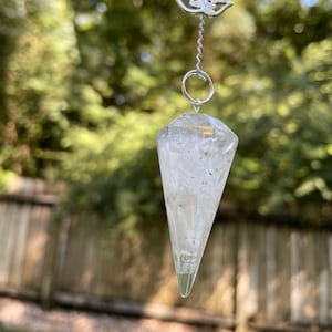 May include: A clear quartz crystal pendulum with a silver chain and a silver lotus charm. The pendulum is hanging in front of a blurred green background.