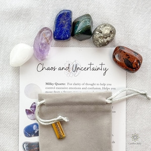Peut inclure: Un ensemble de pierres précieuses polies de différentes couleurs, dont blanc, violet, bleu, vert et marron, disposées autour d'une carte avec l'inscription "Chaos and Uncertainty". Un petit sac gris avec une étiquette dorée "WARRIOR" est également présent.