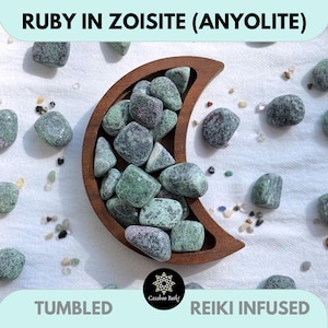 Puede incluir: Un cuenco de madera en forma de media luna lleno de piedras de rubí en zoisita pulidas, también conocidas como anyolita. Las piedras son una mezcla de tonos verdes y morados. El texto "RUBY IN ZOISITE (ANYOLITE)", "TUMBLED" y "REIKI INFUSED" es visible.