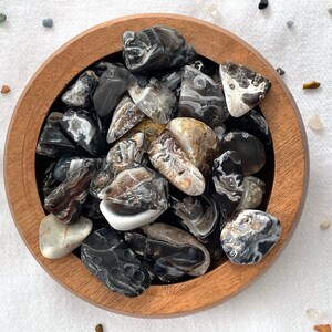 May include: A wooden bowl filled with black and white agate tumbled stones. The stones are smooth and polished, with a variety of patterns and swirls.