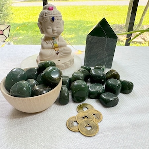 Puede incluir: Una colección de piedras de jade verde en un cuenco de madera, junto con una pequeña estatua de Buda meditando en jade, una pirámide de jade y varias monedas chinas.
