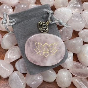 Puede incluir: Una piedra de palma de cuarzo rosa con un diseño de flor de loto dorada. La piedra está en una bolsa de terciopelo gris con un dije de loto dorado.