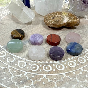 May include: A collection of eight polished gemstone cabochons in various colors, including green, brown, purple, pink, red, white, and blue. The stones are arranged on a wooden tray with a decorative pattern.