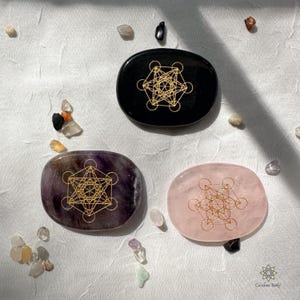 Puede incluir: Tres piedras pulidas de forma ovalada con un diseño geométrico dorado. Las piedras son negras, moradas y rosas.