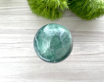 May include: A translucent, faceted fluorite sphere in shades of green and white. The crystal ball is set against a light wood-grain surface, with a backdrop of green foliage. This decorative sphere is ideal for home decor.