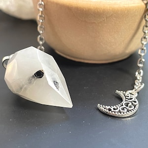May include: White crystal pendulum with a black crystal accent, hanging from a silver chain with a crescent moon charm.