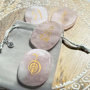 May include: Four pink rose quartz stones with gold symbols, including the symbol for abundance, are displayed on a wooden surface with a decorative pattern. The stones are in a small grey pouch with a silver charm.