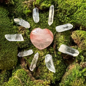 Op de afbeelding: Een roze hartvormige rozenkwartskristal omringd door heldere kwartskristallen op een bed van groen mos.