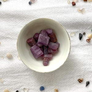 Może przedstawiać: Mała biała miseczka wypełniona fioletowymi i czerwonymi kamieniami szlachetnymi. Miseczka stoi na białym tle z tkaniny.