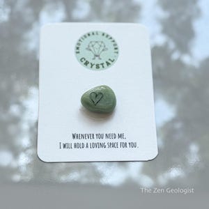 Puede incluir: Un cristal verde con un símbolo de corazón negro, sobre una tarjeta blanca. La tarjeta tiene un logotipo circular que dice "Emotional Support Crystal". El texto en la tarjeta dice "Whenever you need me, I will hold a loving space for you."