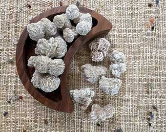 May include: A collection of white selenite rose quartz crystals in a wooden crescent moon-shaped bowl. The crystals are arranged in a decorative pattern and are surrounded by a beige textured background.
