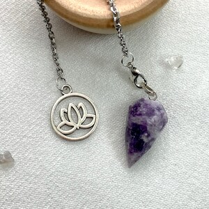 May include: A purple amethyst crystal pendulum with a silver chain and a silver charm featuring a lotus flower design.