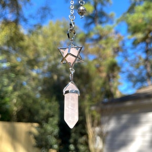 Puede incluir: Un péndulo de plata con una punta de cristal de cuarzo rosa y un cristal de cuarzo rosa enjaulado en forma de estrella de David.