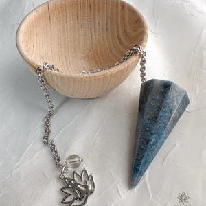 May include: A blue and grey crystal pendulum with a silver chain and a silver lotus charm. The pendulum is hanging over a wooden bowl.