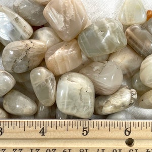 May include: A collection of white and light brown moonstone tumbled stones. The stones are approximately 1 inch in size and are arranged on a white fabric background. A wooden ruler is placed below the stones for size reference.