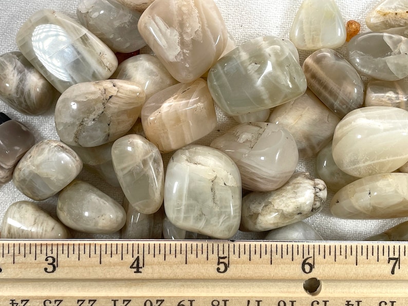May include: A collection of white and light brown moonstone tumbled stones. The stones are approximately 1 inch in size and are arranged on a white fabric background. A wooden ruler is placed below the stones for size reference.